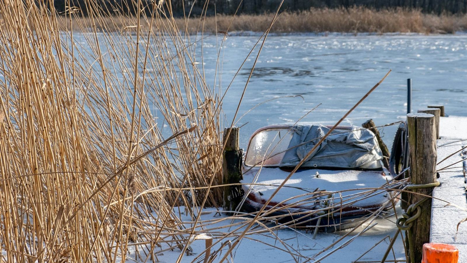 Covered boat locked in ice beside a dock on a frozen lake — winterize the engine before the first hard freeze catches it in the water
